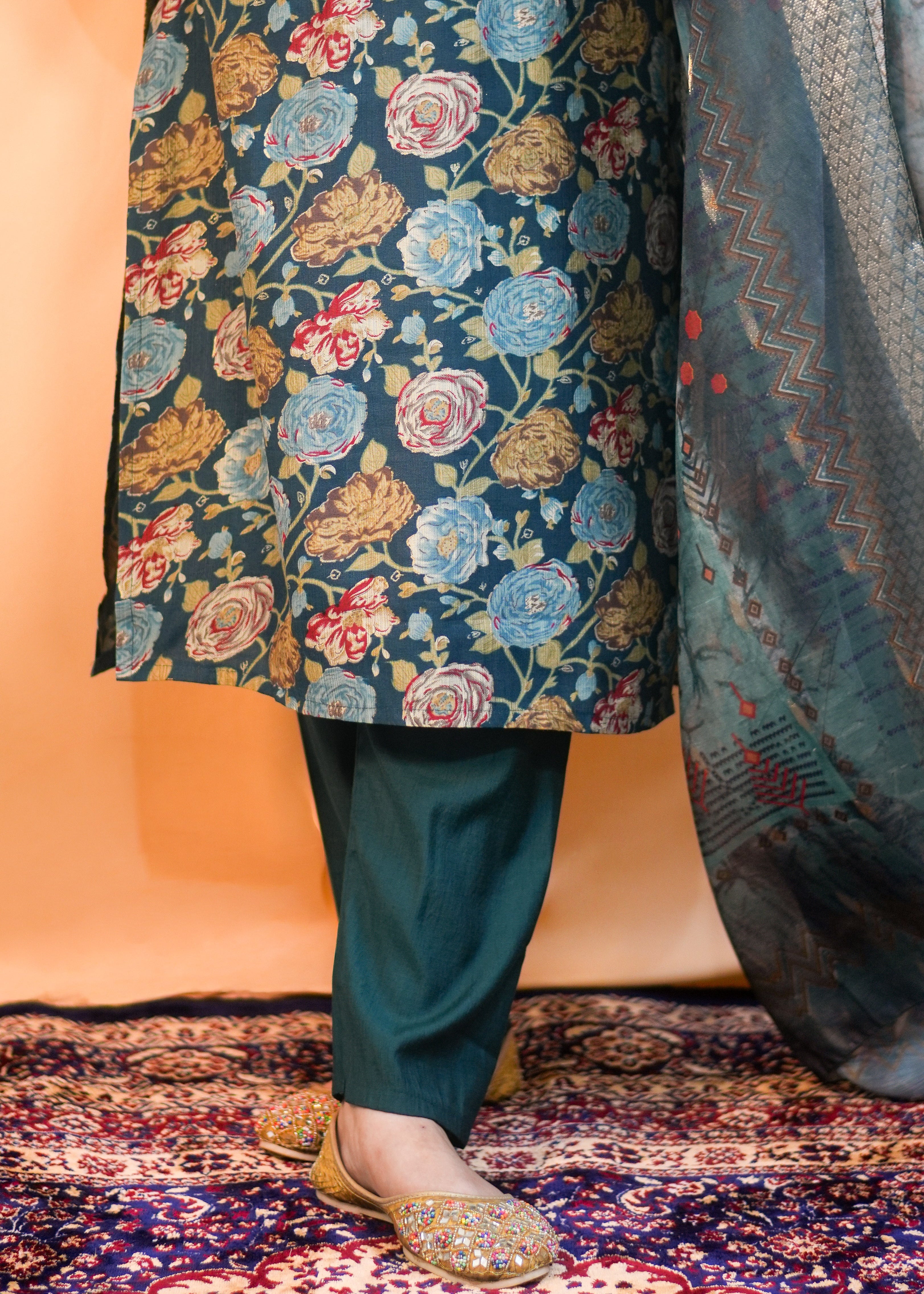 Person wearing a floral-patterned long dress with matching pants on a patterned carpet.
