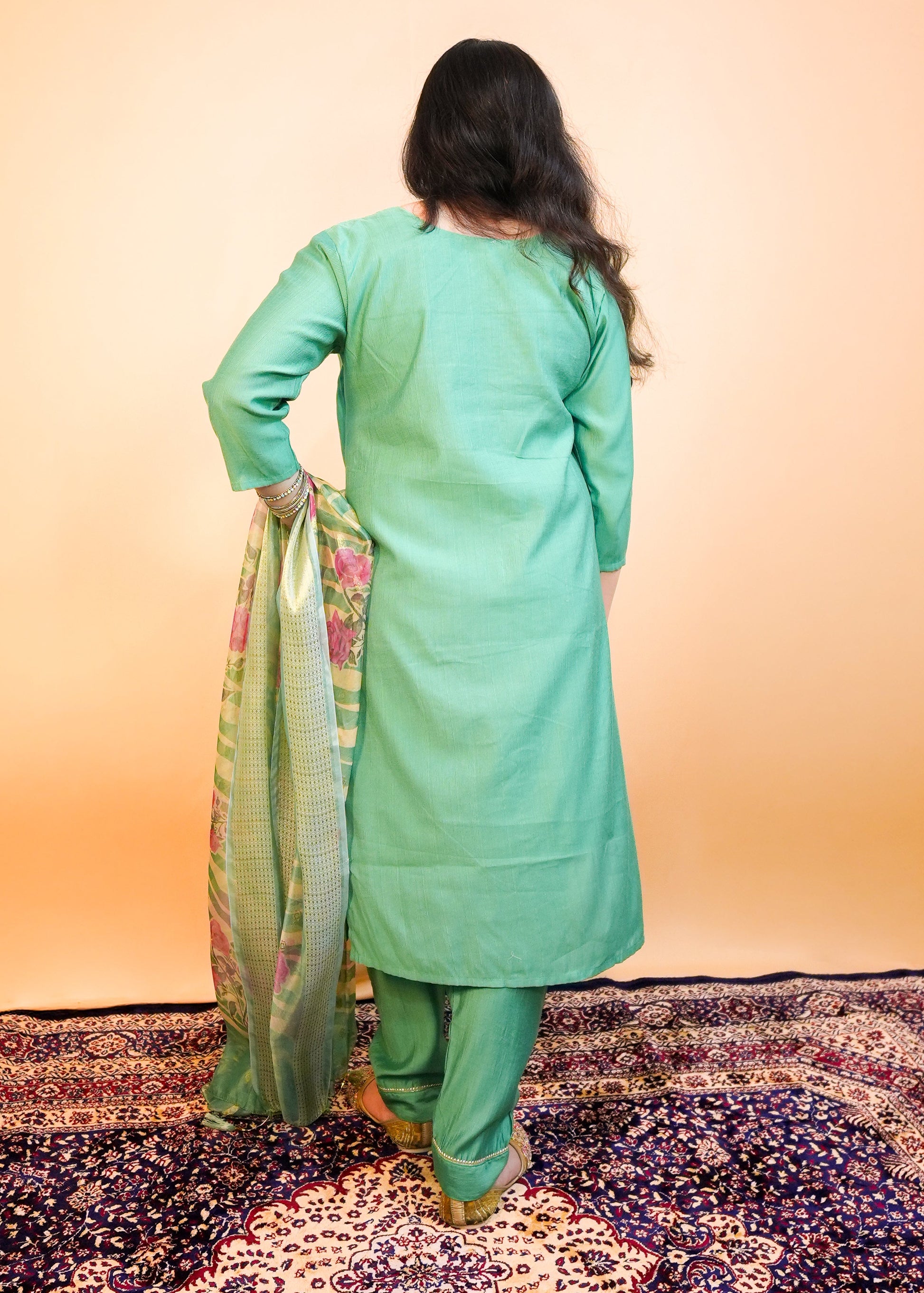 Woman in a green traditional outfit standing on a patterned rug with a plain background