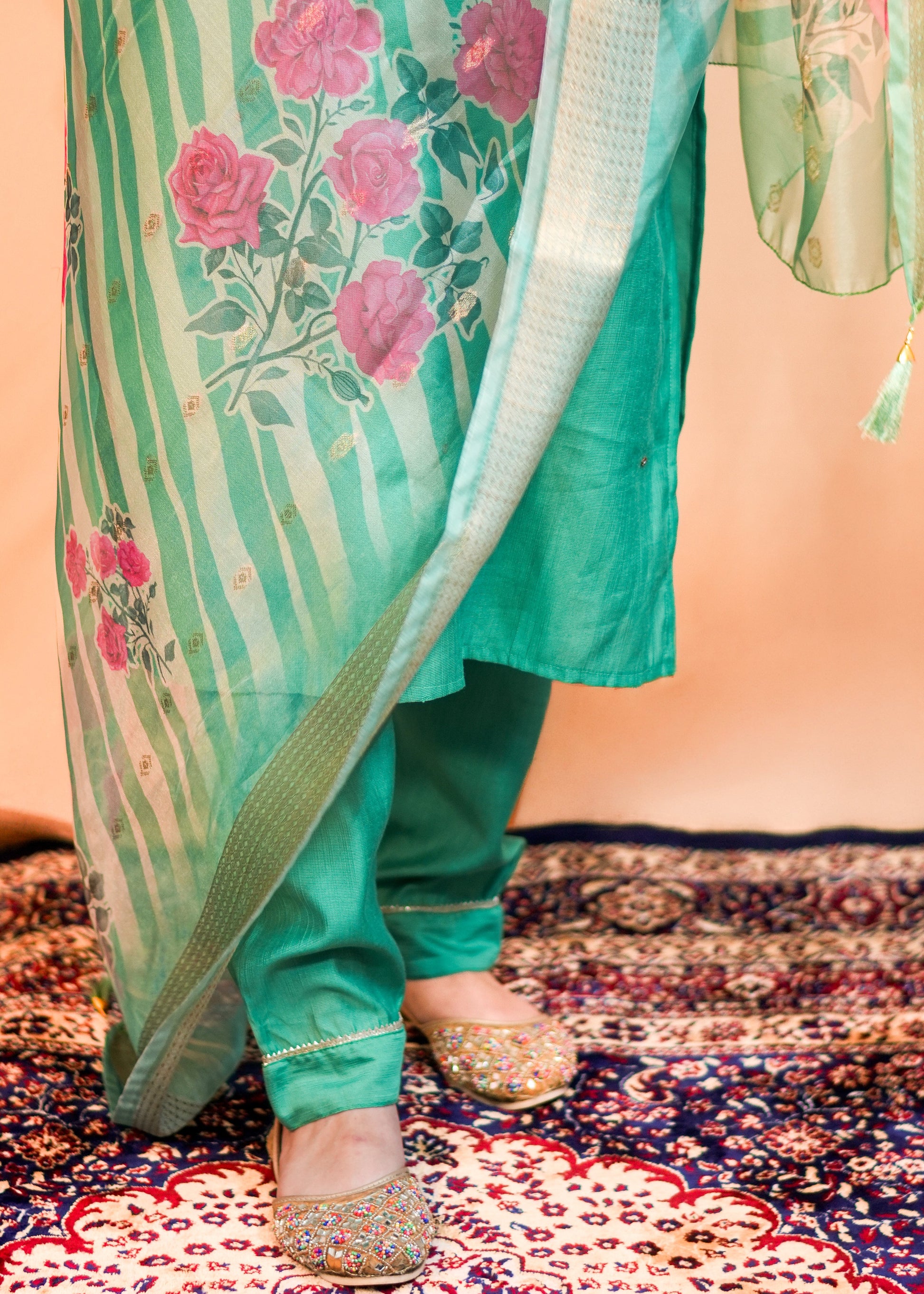 Person wearing a green traditional outfit with floral patterns on a patterned rug.