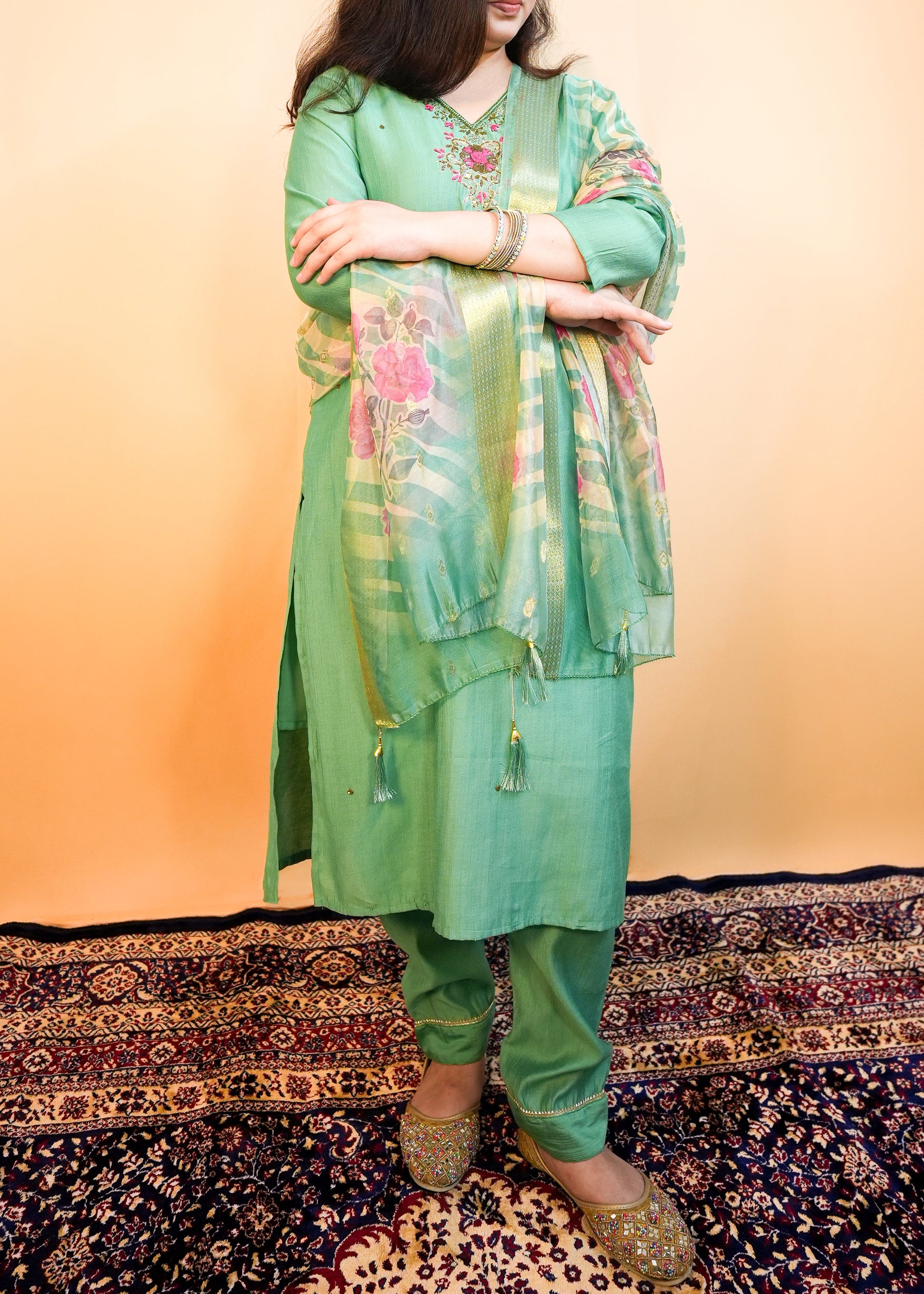 Woman wearing a green traditional outfit with floral patterns on a patterned rug.