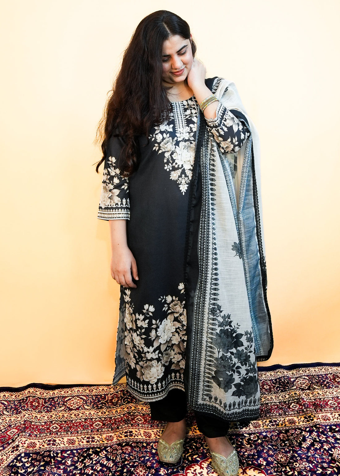 Woman in traditional black and white outfit with floral patterns on a patterned rug.