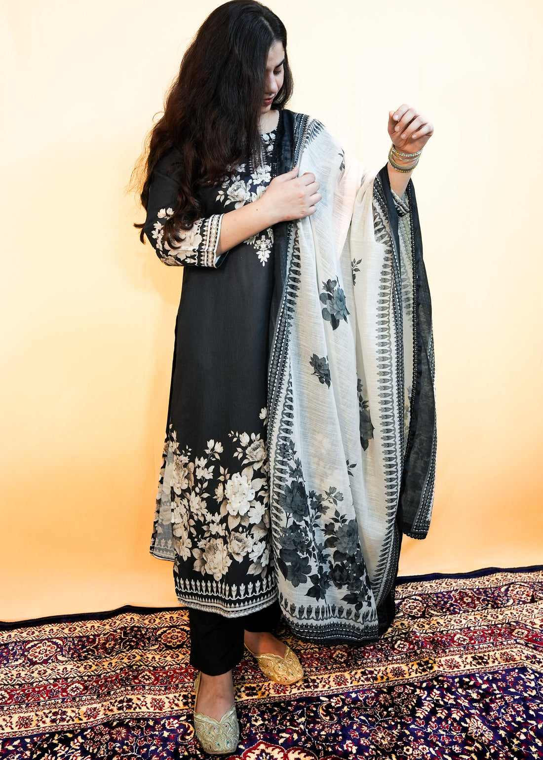 Woman in traditional black and white outfit with floral patterns on a patterned rug.