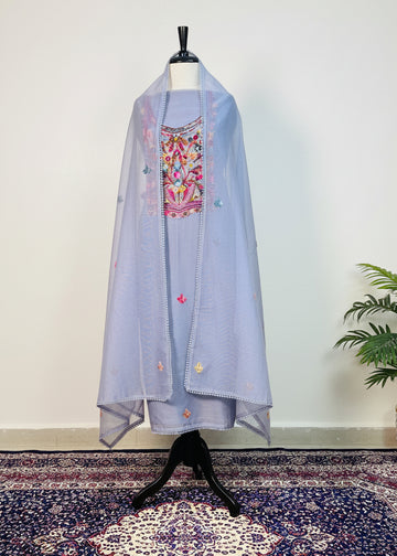 Light blue embroidered garment on a mannequin against a white wall with a plant in the corner.