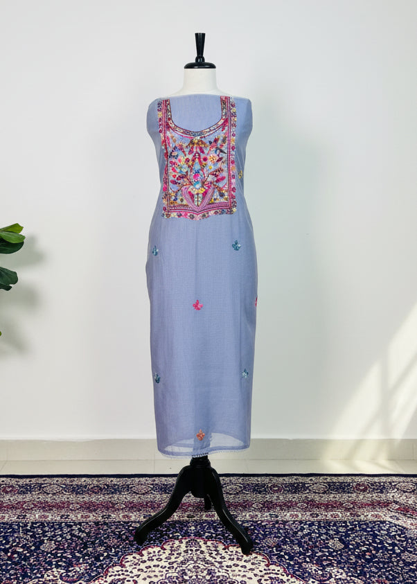 Light blue embroidered garment on a mannequin against a white wall with a plant in the corner.