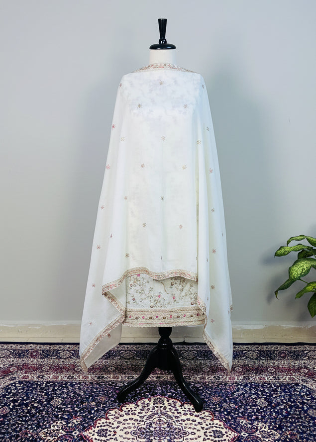 White embroidered garment on a mannequin in a room with wooden floor and wall lights.