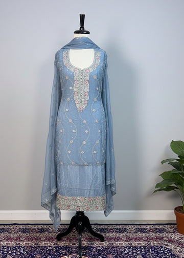 Mannequin wearing a light blue embroidered outfit on a patterned rug with a beige curtain background